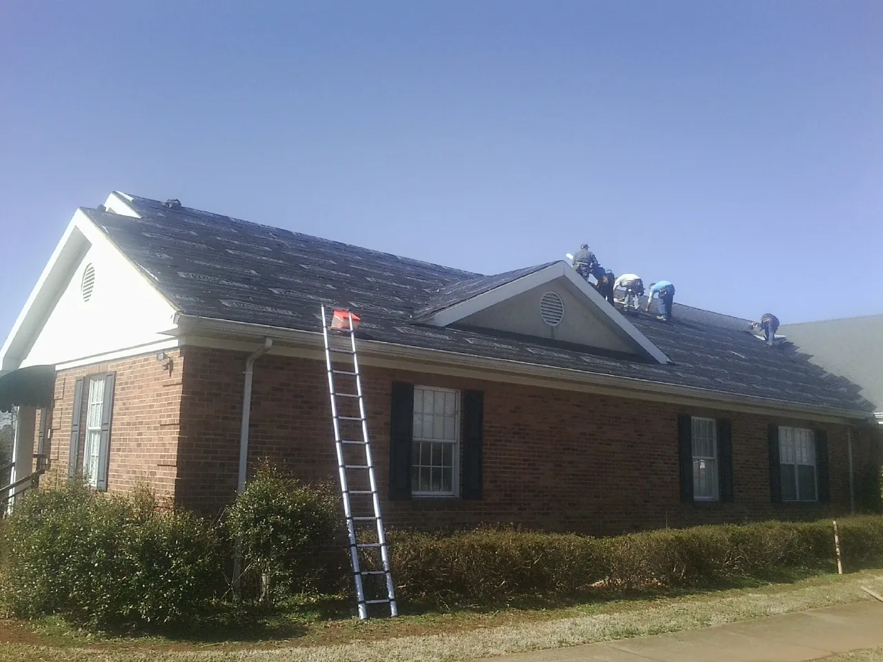 Crew tearing shingles off a brick ranch roof with a ladder set — Emergency Roof Repair work by our crew in Gibbon
