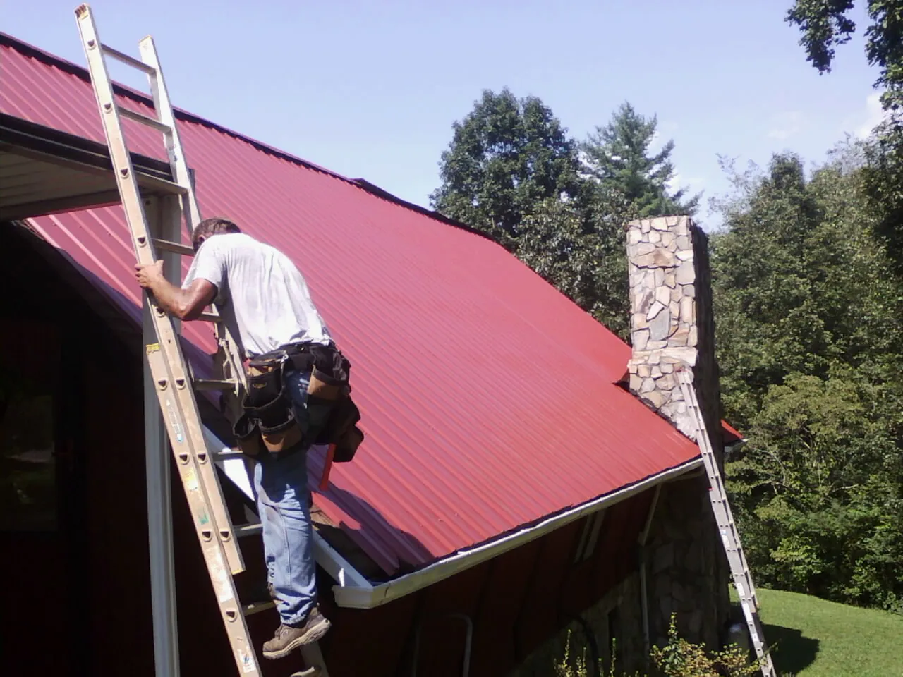 Roofer on a ladder beside a red metal roof and stone chimney — Soffit & Fascia Repair by Roofing Brothers in Gibbon
